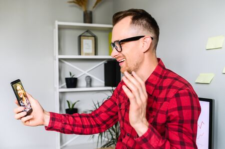 Smiling guy is making a video call via phone, he sits at the workplace and waving hi to his female colleague , a shelf with plants and decor elements on it is on the backgroundの写真素材