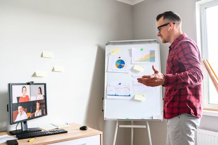 Confident young man conducts webinar, business training online. Online coach stands near flip chart in front of PC display and explains something to online audienceの写真素材