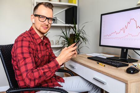 A young man in glasses sits in front of the computer with graphs on the screen at the office desk, he is holding phone and looks into cameraの写真素材
