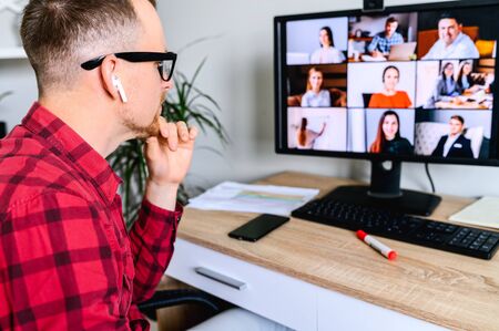 Online video conference. Young man connects via video call with employees, he sits at the office desk, touching his chin with a hand, wearing glasses . App for video chat with employeesの写真素材