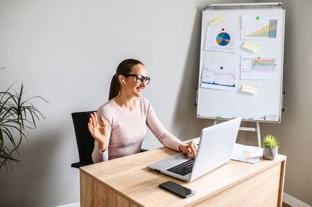 Woman in office has video conversation. Young positive woman in glasses sittung at the desk in front of laptop and talking with somebody online, flip chart on backgroundの写真素材