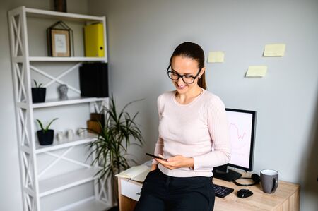 Smiling woman looks at the phone screen and standing in home office, plants and computer on the background. Business woman is watching something on the phone while leaning on office desk.の写真素材