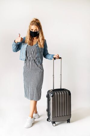 Girl in face mask standing with suitcase isolated. Young woman in face mask showing thumb up and stands with a flight bag in studio with a white background.の写真素材