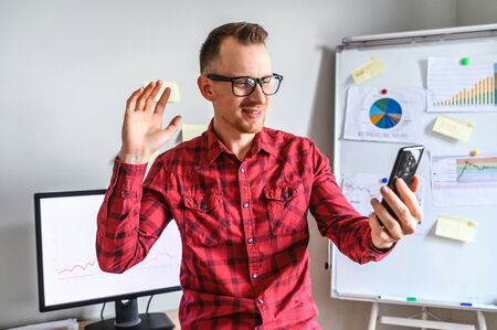 Smiling hipsterich guy in glasses is using his phone to make a video call and waving hi into phone camera, he stands in the office, a workplace with a PC and flipchart on the backgroundの写真素材