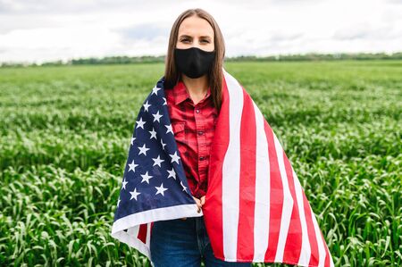 Sad girl wearing face mask stands among green field wrapped herself in a USA flag, she looks at camera seriouslyの写真素材