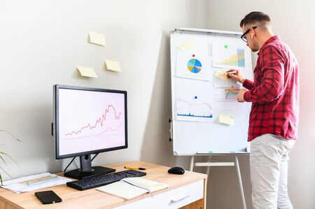 Guy in casual plaid shirt stands in office, he is making notes on a flipchart, PC monitor on the office desk on the background. Data analysis in processの写真素材