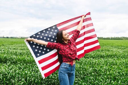 Cheerful beautiful young woman running through green field with her hands spread up and holding blowing american flag. Independence day, fourth julyの写真素材