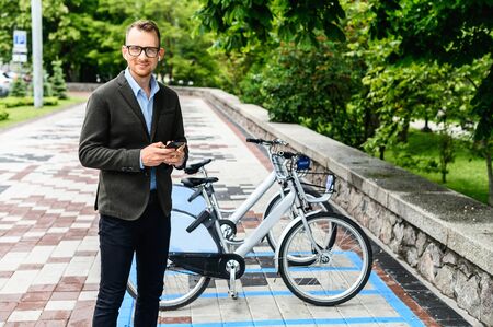 A young businessman in smart casual using app for rent city bike. A guy wearing glasses is typing on the smartphone, parking of city bicycles on the backgroundの写真素材