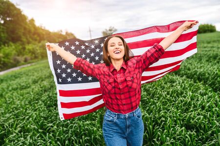 Happy attractive young woman is walking through green field, her hands raised up with US flag. Freedom and democracyの写真素材
