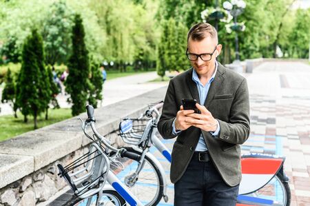 A young businessman in smart casual using app for rent city bike. A guy wearing glasses is typing on the smartphone, parking of city bicycles on the backgroundの写真素材