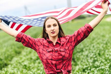 Attractive young woman is walking through green field, her hands raised up with flying US flagの写真素材