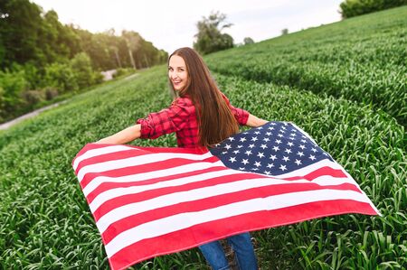 Carefree attractive young woman is running through green wheat field and laughing, her hands up with flying USA flagの写真素材