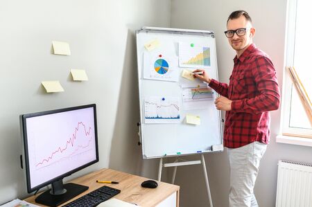A smiling affable guy in casual plaid shirt stands in office, he is making notes on a flipchart and looks in camera, PC monitor on office desk on the backgroundの写真素材