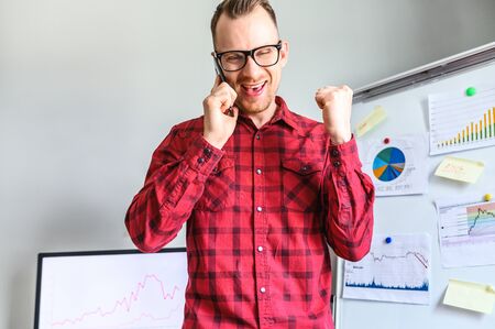 Satisfied young man in glasses is rejoicing and smiling while talking on phone, his arm bent up at the elbow, he struck a deal over the phone. PC screen and flipchart on the backgroundの写真素材