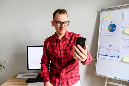 Cheerful guy in glasses talks on the phone using a video frontal camera, he holds his phone aloft, he sits leaning on office desk, PC mpnitor and flipchart on backgroundの写真素材