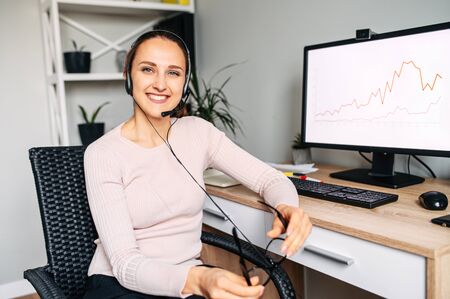 Friendly office atmosphere. Positive young woman in glasses works with PC and using headset indoor. She looks at camera with smileの写真素材