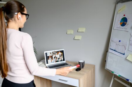 Online presentation. Smart woman in casual wear with glasses talks speech in front of online audience on laptop screen. Woman points on flip chart. Back viewの写真素材