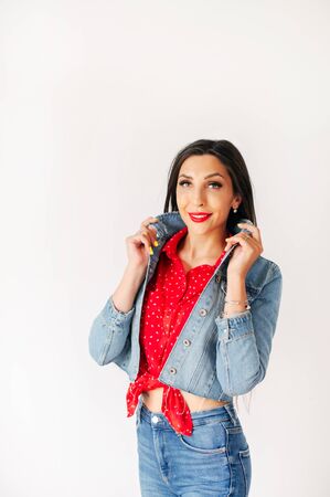 Lovely young brunette woman in jeans suit and red shirt posing on white background. Vertical shotの写真素材
