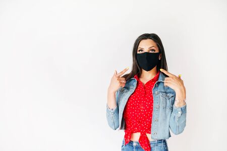Girl stands on white background in black protective mask on the face points fingers on the maskの写真素材