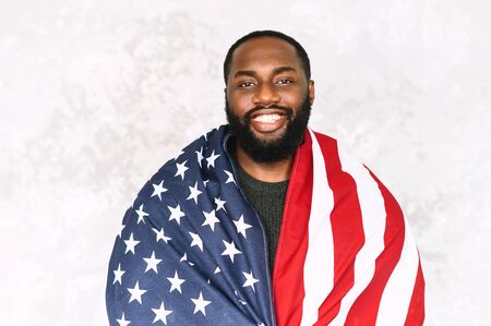 Happy African-American guy wraps himself in the U.S. flag and looks at the camera and laughs. Studio portrait close-upの写真素材