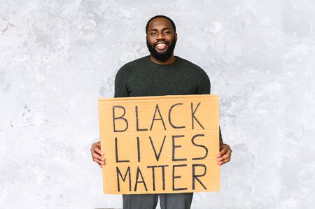 Handsome black guy stands and smiles on a gray background, he is holding board with inscription black lives matter. Studio shotの写真素材