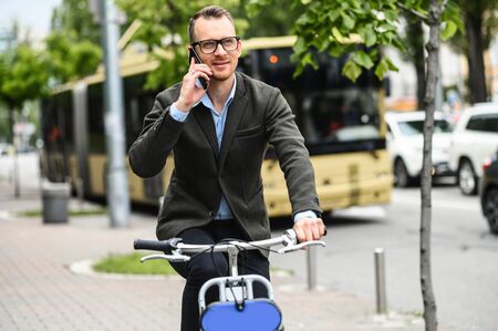Use environmentally friendly modes of transport. Cheerful young man in smart casual wear and eyeglasses is riding a bike and talking on the phone while getting to the officeの写真素材