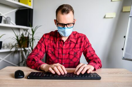 A web camera shot of a busy young office worker typing on the keyboard, he wears mask in order to protect his colleague. Work while quarantineの写真素材