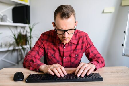 Webcam view of young concentrated man in casual plaid shirt at the office, he is typing on the keyboard and looking on it with a serious look on his face.の写真素材
