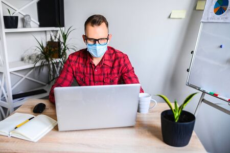 A young business man works at the office, he sits with a laptop at the office desk wearing a medical mask. A mid shot of a guy working in mask, houseplant and flip chart on the backgroundの写真素材