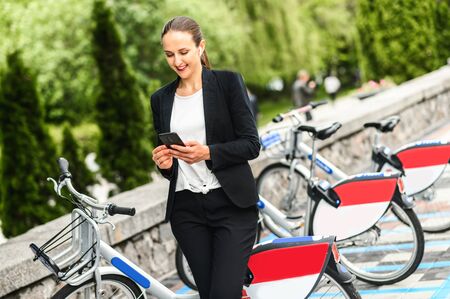 Use environmentally friendly modes of transport. Young woman is using app on the phone for renting a city bike, she stands with a smartphone near parking of bicycleの写真素材