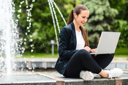 Female student sits near city fountain in the park and is using laptop for preparing homework. An attractive young girl sits in a relaxed position, typing on the keyboard and smilingの写真素材