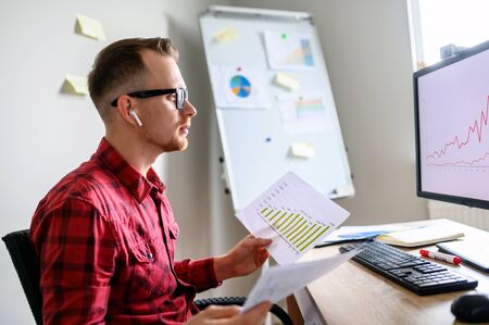 Young man is checking different statistics and comparing it to an information on computer. He sits in front of PC beside the window and holds papers, flipchart and workplace on backgroundの写真素材