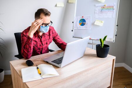 Pensive young worker sits in front of the laptop, working in medical mask at the office, he is thinking over something and leaning his head on a handの写真素材