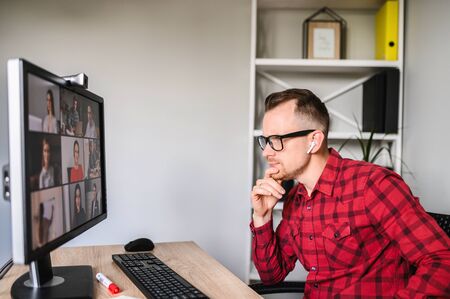 Pensive young bearded man talks over an app on PC via video call with his colleagues. He sits in...