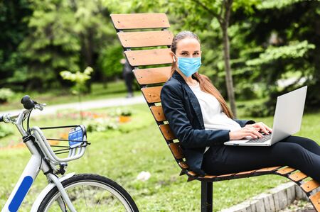 Young confident woman in a medical face mask sits on the bench outdoors is using laptop for answer emails, city bike in near. Precautions for epidemics, allergies, smogの写真素材