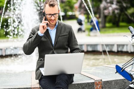 Successful young businessman wearing eyeglasses and smart casual clothes talks on the phone using laptop for work outdoorsの写真素材