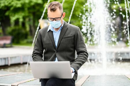 A young guy wearing medical face mask is working with laptop outdoors in the city, he sits near city fountain in the square. Protect from viruses, allergyの写真素材