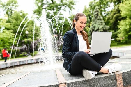 Female student sits near city fountain in the park and is using laptop for preparing homework. An attractive young girl sits in a relaxed position, typing on the keyboard and smilingの写真素材