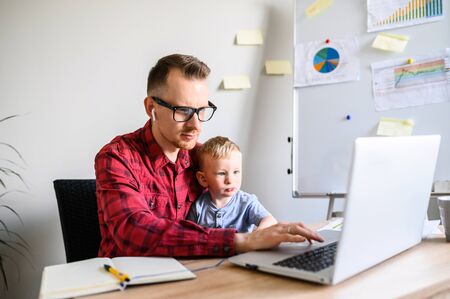 Father works from home office while babysitting with toddler kid. Dad sits at the desk with a little son on his laps and typing on keyboardの写真素材