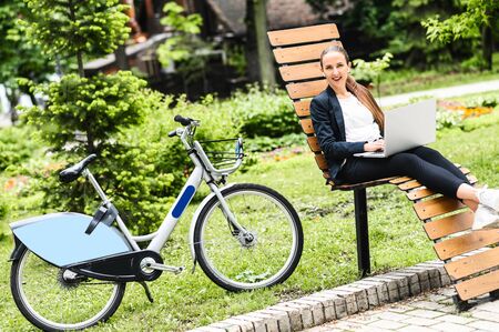 Young modern business woman cycled to the park to work with a laptop outdoors. Woman in smart casual wear sits with a laptop on the bench near bike and looks at cameraの写真素材