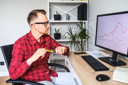 A young thoughtful man looks at the PC screen with graphs on it, he sits at the office desk and holds a pencil wearing glassesの写真素材