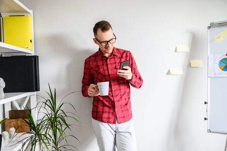 Relax office employee is having a cup of coffee while his dinner break, he stands in the office and checking his phone, Flipchart and office shelf on the backgroundの写真素材
