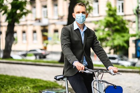 Young guy with a medical mask on the face in smart casual wear is riding a bicycle in the city. Precautions for epidemics, allergies, smogの写真素材
