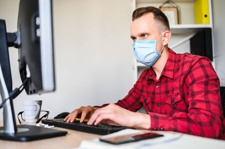 Responsible employee works with the computer wearing a protective mask. A young man works in the office while quarantine.の写真素材