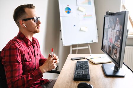 Online meeting of a young man with his subordinates. He sits accross from the window talking via web camera on PC to a group of people at the workplace, flipchart on the backgroundの写真素材