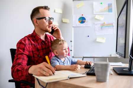 A young man works from home while quarantine, he sits at the computer and talks on the phone, his kid sits on his lap. Young dad works in home office with a kid in front of a PCの写真素材