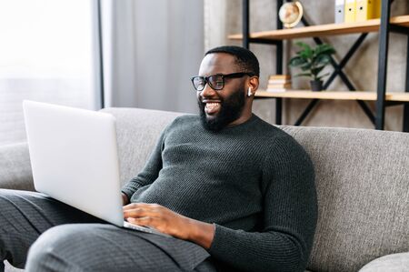 Happy African-American guy sits on sofa in modern living room alone and using trendy laptop for net searching, web surfing or texting messagesの写真素材