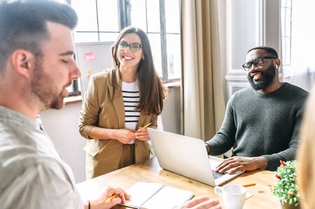 A friendly atmosphere in the office. Young, confident employees of the multiracial team discuss tasks and laugh at the morning meetingの写真素材