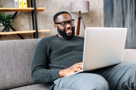 Cheerful African-American guy sits on sofa in modern living room alone and using trendy laptop for net searching, web surfing or online learningの写真素材