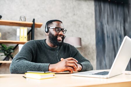 Young African-American guy is call center worker or support. Smart black man in eyeglasses uses a handsfree headset and laptop to talk online at his workplaceの写真素材